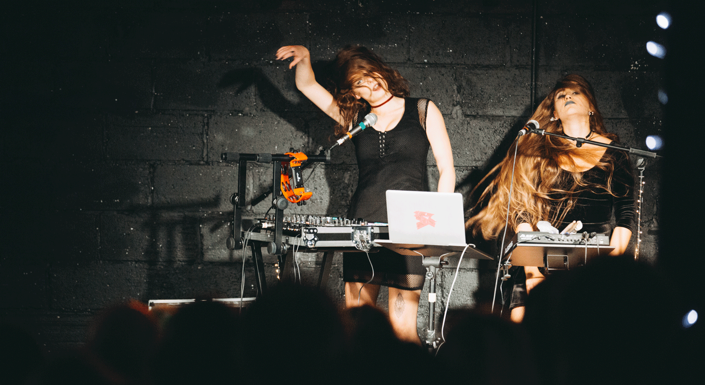 Two women in black dresses play electronic instruments on stage, shaking their heads and hair