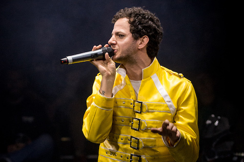 A man in a yellow Freddie Mercury-style jacket sings into a handheld microphone