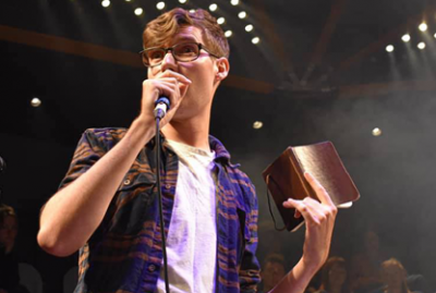 A man in white t-shirt and plaid shirt reads from a notebook into a mic