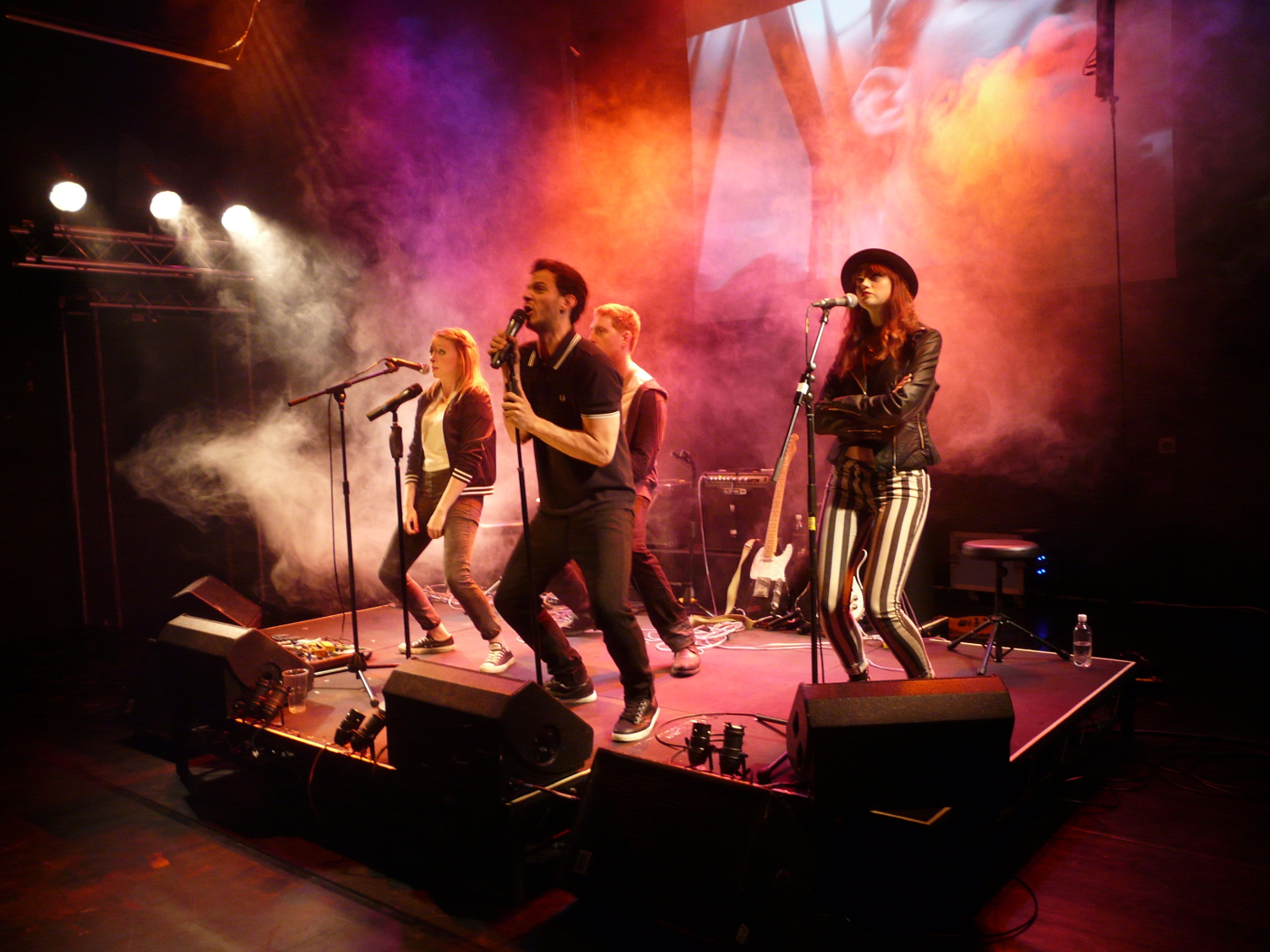 Four performers on stage, posed as a band, singing into microphones, bathed in pink-red light
