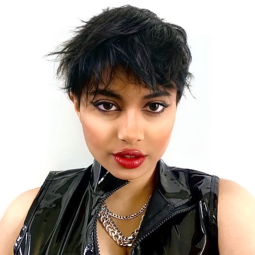 Headshot of androgynous Indian person wearing a latex vest top, silver chains and red lipstick.