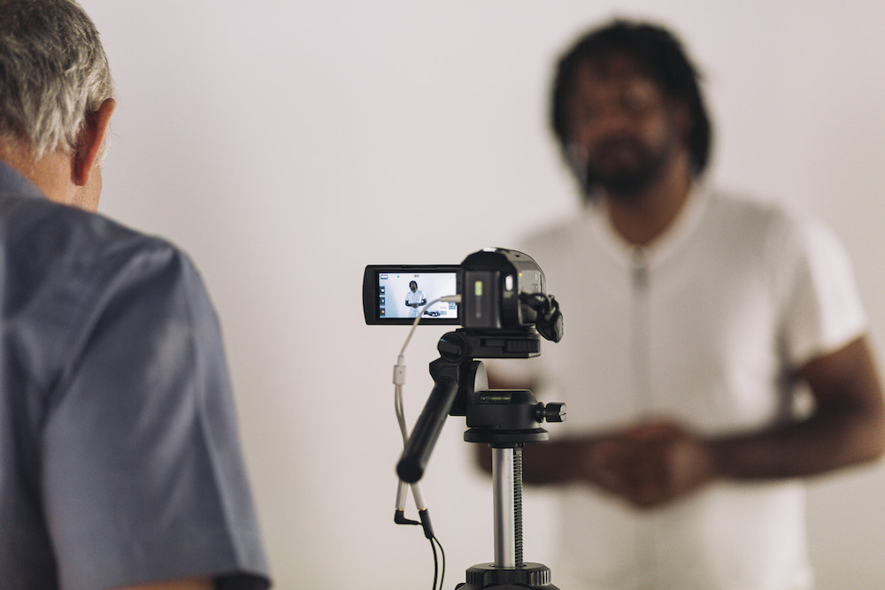 A video camera pointing at a tall Black man in a white t-shirt