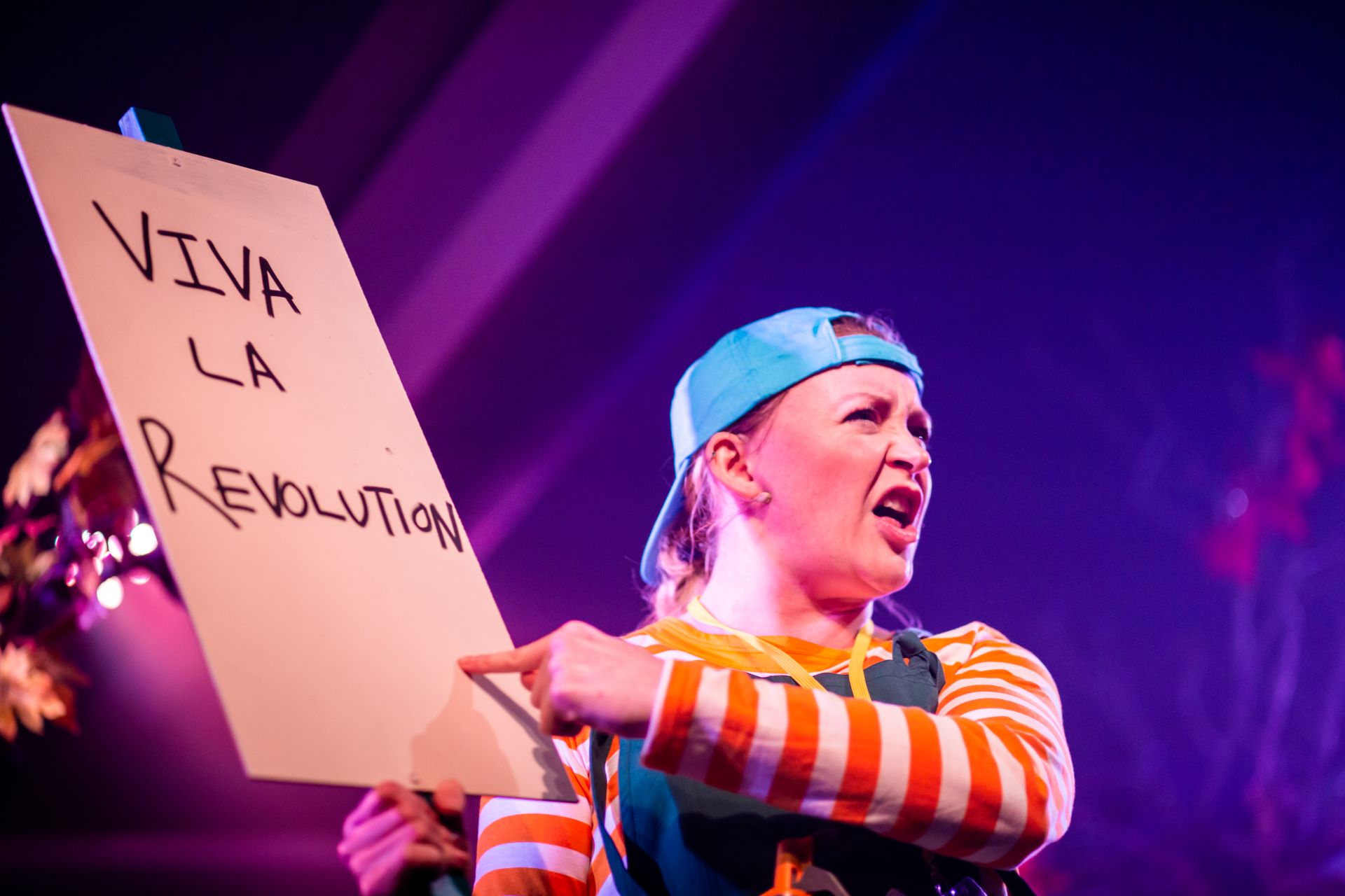 A white woman in stripey top, dungarees and backwards baseball cap holds a placard that says 'viva la revolution'