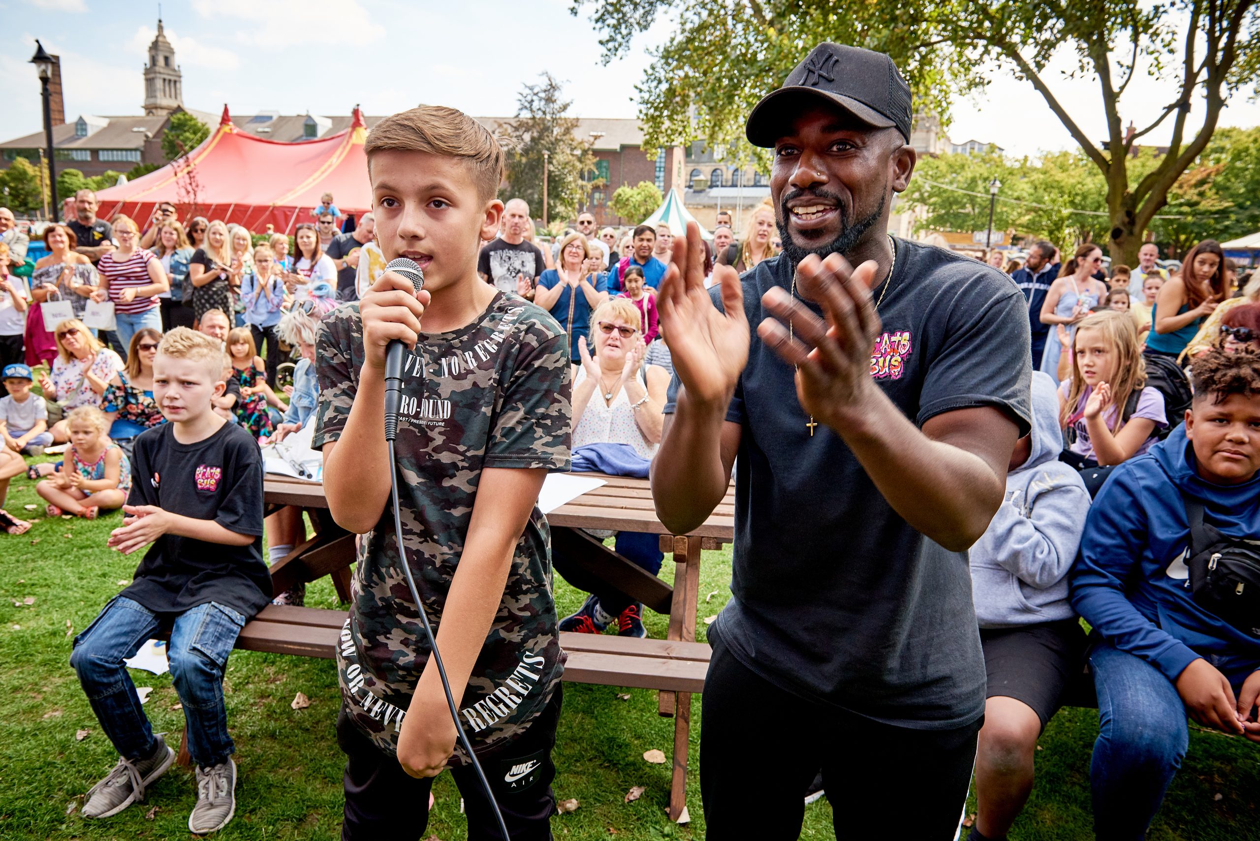 A Black man applauds a white boy on a microphone. In the background lots of children watch.