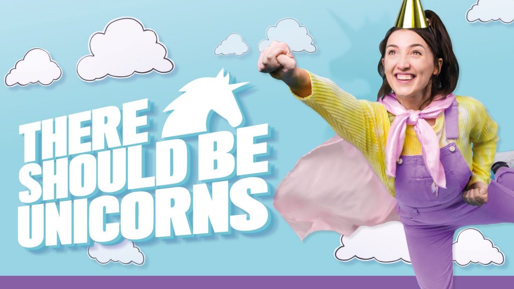 A white woman with long dark hair, in lilac dungarees, yellow jumper, gold party hat and pink cape makes a superhero pose, against blue background with white clouds and text that says "There Should Be Unicorns"