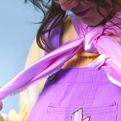 A white woman with long dark hair, in lilac dungarees, yellow jumper holds a pink cape