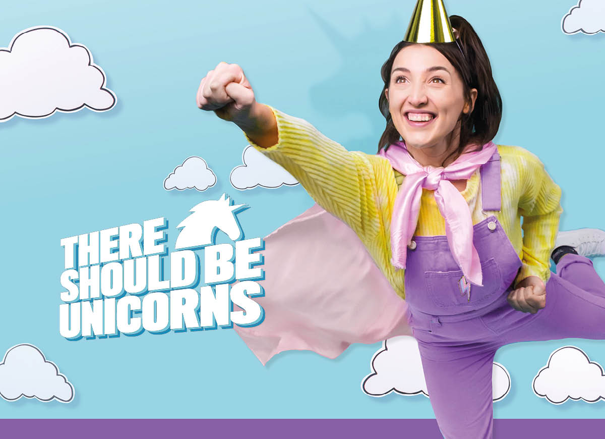 A white woman with long dark hair, in lilac dungarees, yellow jumper, gold party hat and pink cape makes a superhero pose, against blue background with white clouds and text that says 