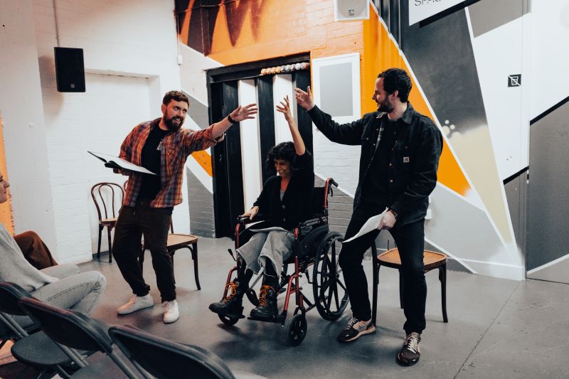 An Asian woman in a wheelchair and two white men, all holding scripts, make the cheers gesture
