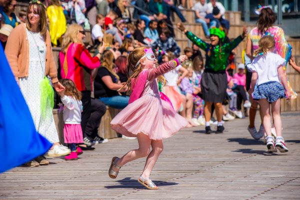 A young child in a pink dress, pink cape and pink and silver unicorn hero mask is punching her fist into the sky and kicking her leg back into a flying superhero pose.