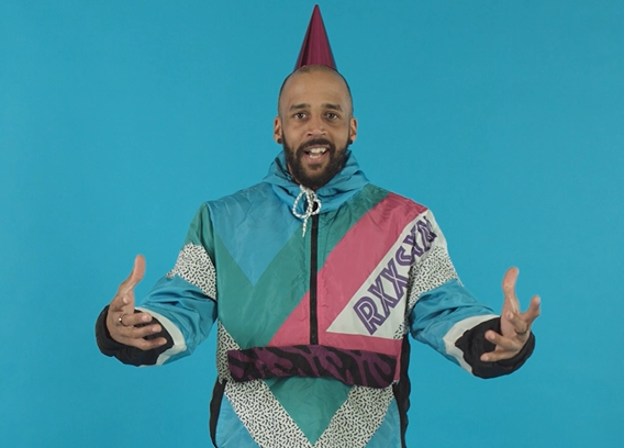 A mixed race Black man in ski-style jacket and pink party hat speaks to camera