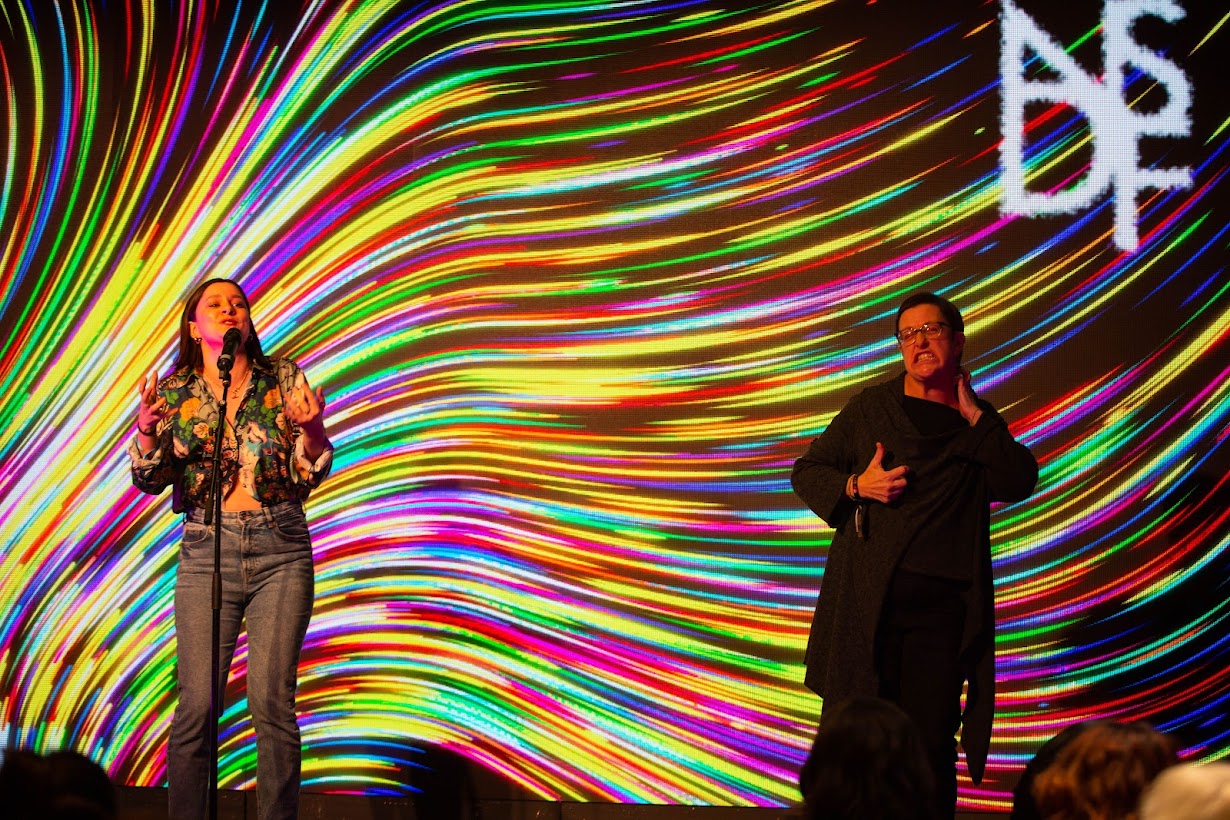Two performers on stage in front of a multicoloured kaleidoscopic background.