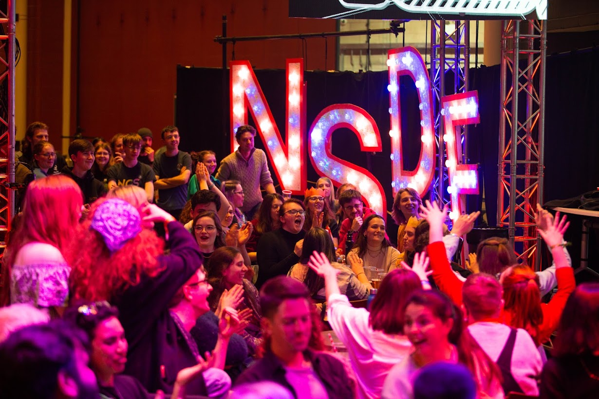 An excitable seated audience bathed in pink-red light, in front of giant lit-up letters that spell NSDF