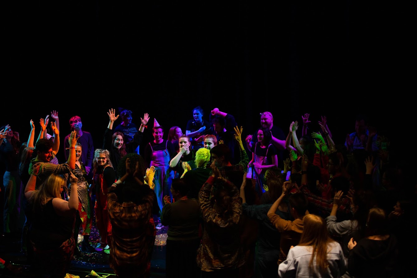 An audience invades the stage to dance with their hands in the air under various coloured stage lights