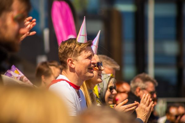 A white man in a red cape, white top and holographic pink triangle party hat sits in a crowd smiling.