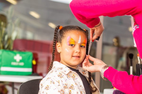 A young child with two braids in her hair is having her face painted with a unicorn horn.