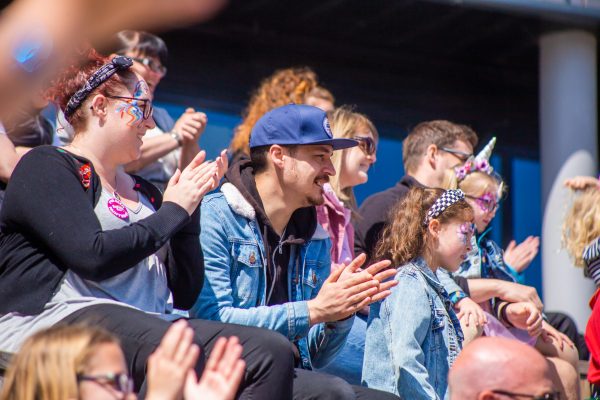 A white man with dark facial hair and a blue cap claps whilst sat in the audience next to a woman with red hair and a young child in a denim jacket with dark hair.