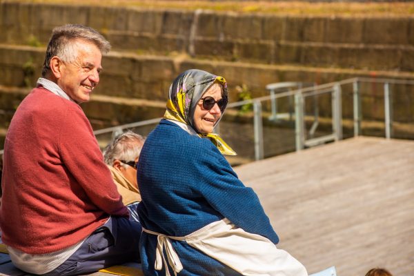 An older man and woman smile at the camera over their shoulders. The man is on the left wearing a red jumper over a white shirt and blue trousers. The woman is central and is wearing a blue top, a grey and yellow headscarf, a cream apron and black sunglasses.