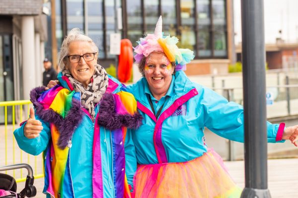 Two older white women are dressed in bright blue jackets and smiling at the camera. The woman on the left has a rainbow cape and the woman on the right has a rainbow tutu and rainbow unicorn headband.