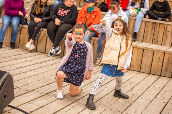 Two children make superhero poses for the camera. The child on the left has rainbow face paint and is down on one knee with one arm raised in the air. The child on the right has dalmatian face paint and is stood with one arm raised in the air.