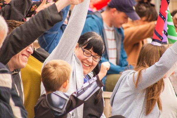 A white woman with black hair and glasses smiles at a young child next to her sitting in a crowd. They both have have one arm in the air making a superhero pose, as do the crowd around them.