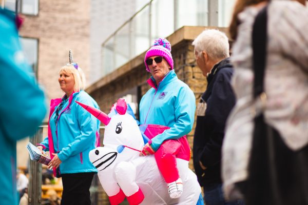 A white man in a bright blue jacket, a pink and purple bobble hat, and a wearable inflatable unicorn.