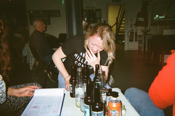 A woman laughs in front of a table with a quiz sheet and bottles on.