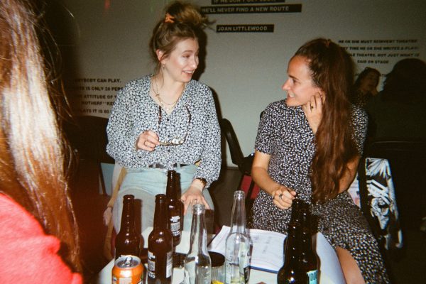 Two women are laughing with each other in front of a table full of beer bottles.