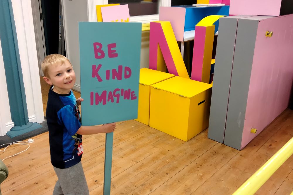 A small boy holds a sign that says "Be kind imagine" from the set of There Should Be Unicorns