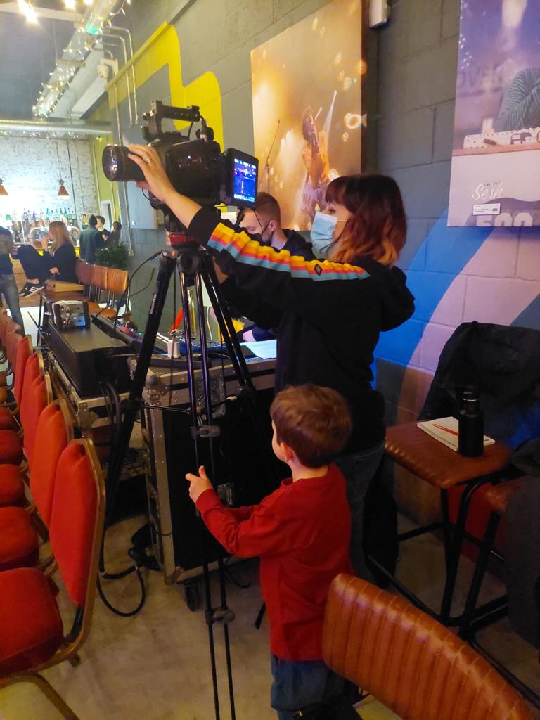 A small boy stands next to his mum, who is operating a camera on a tripod