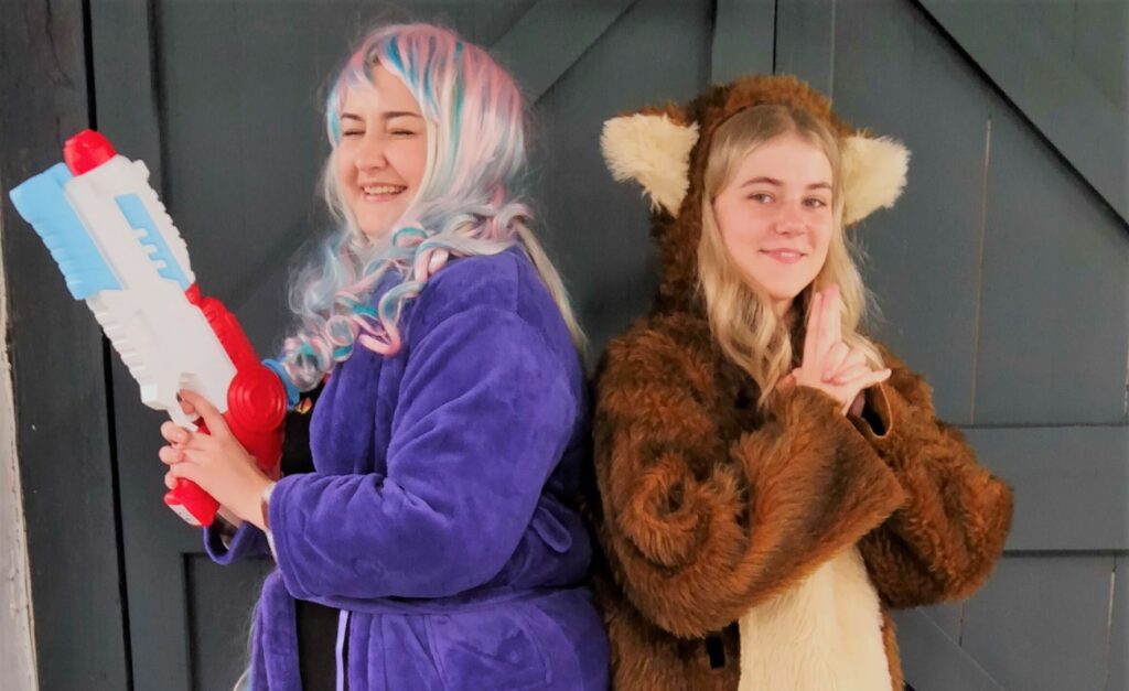 A young white woman in purple bathrobe and wig, holding a water pistol, stood back to back with another young woman dressed in a fox onesie