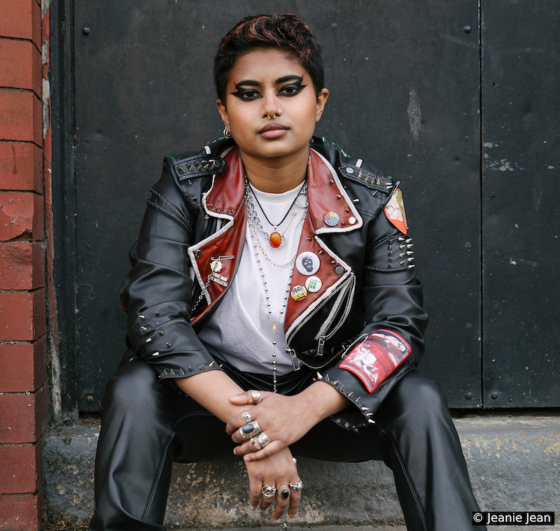 Jay Mitra, a South Asian nonbinary person, is sat on the steps outside looking directly at the camera. They have black hair and are wearing a white t-shirt, a studded black and red leather jacket, and black leather trousers. The photographer Jeanie Jean is credited on the bottom right of the photo.