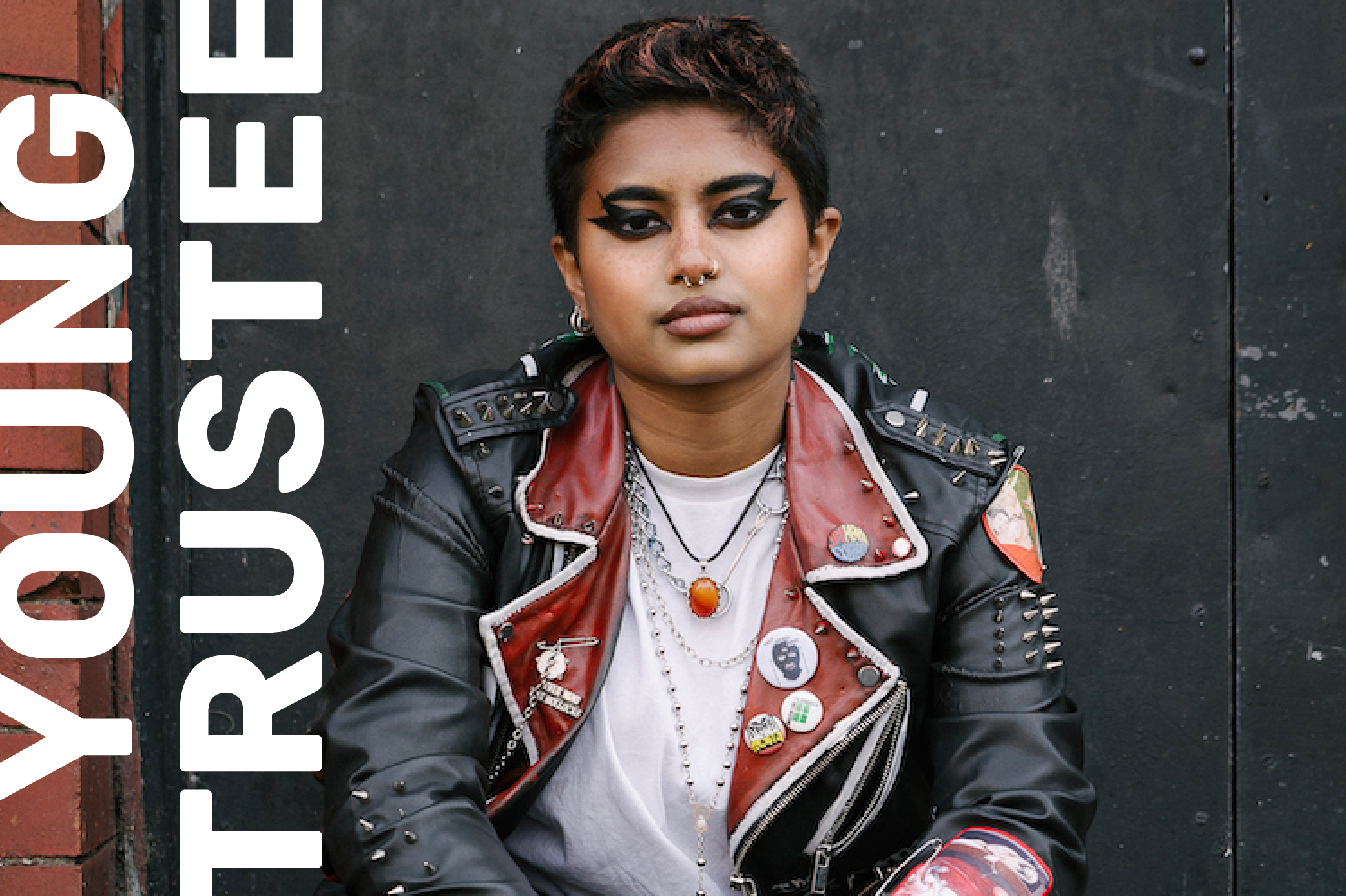 Jay Mitra, a South Asian nonbinary person, is sat on the steps outside looking directly at the camera. They have black hair and are wearing a white t-shirt, a studded black and red leather jacket, and black leather trousers. The photographer Jeanie Jean is credited on the bottom right of the photo.