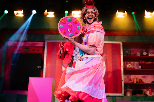 A white man with dark facial hair is dressed as a panto dame in pink dress, red glasses and a cherry hat, holding up a cake.