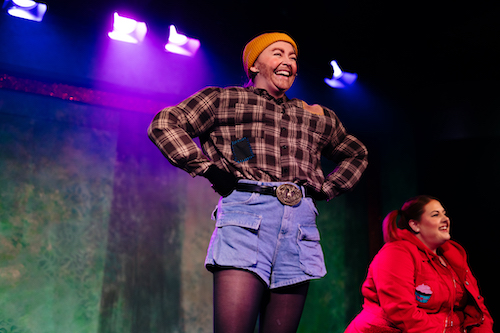 A white woman in drag as a male lumberjack, in a check brown shirt, yellow beanie and denim shorts.