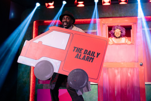 A Black man dressed in a biege overcoat and brown hat holds a 2D cut out of a red van that says The Daily Alarm on the side.