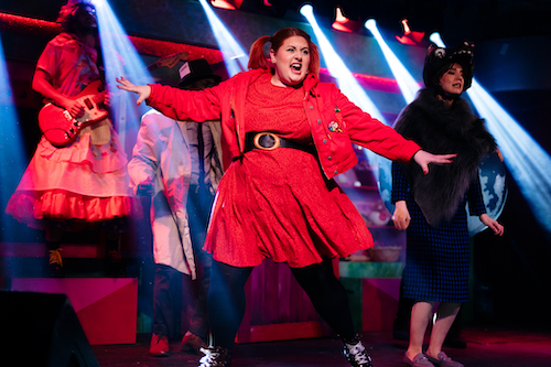 A fat white woman with red hair in bunches dressed as Red Riding Hood dances with arms outstretched. In the background a panto dame plays guitar.
